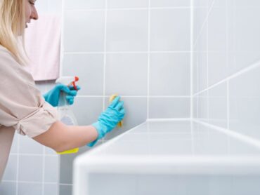 Dona bionda che pulisce mattonelle del bagno, guanti azzurri e detersivo spray in mano